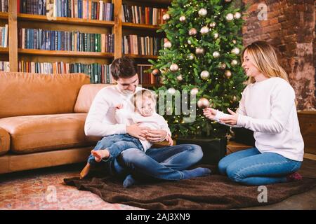 Natale in famiglia concetto. Bella famiglia siede vicino a albero di Natale di sera d'inverno. La famiglia felice con il figlio sul pavimento vicino al divano di casa. La famiglia a casa Foto Stock