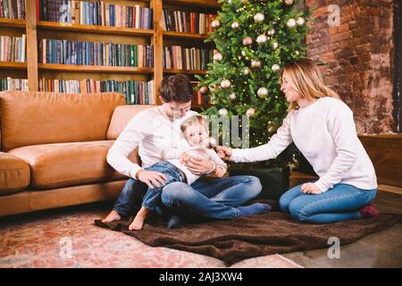 Natale in famiglia concetto. Bella famiglia siede vicino a albero di Natale di sera d'inverno. La famiglia felice con il figlio sul pavimento vicino al divano di casa. La famiglia a casa Foto Stock