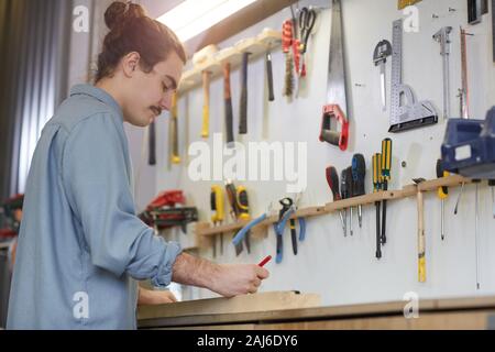 Giovane lavoratore in abbigliamento casual permanente al tavolo e scrivere qualcosa con gli strumenti di lavoro sulle pareti in officina Foto Stock
