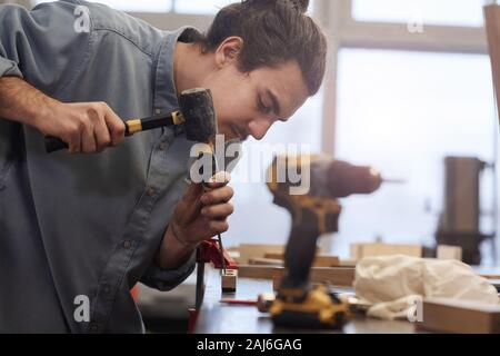 Giovani repairman usando un martello e cacciavite nel suo lavoro al tavolo in officina Foto Stock