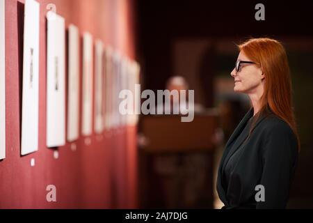 Vista laterale del rosso giovane donna dai capelli in occhiali in piedi di fronte alla parete con dipinti e godendo della tecnica Foto Stock