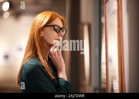 Rosso giovane donna dai capelli in occhiali guardando la pittura e godendo della tecnica nella galleria Foto Stock