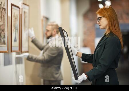 Vista laterale di dai capelli rossi ragazza in occhiali esaminando la pittura nella sua mano e sceglierne uno per se stessa con uomo al lavoro Foto Stock