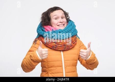 Carino affascinante adolescente di sesso femminile in vestiti caldi, sciarpa e cappello che mostra due pollici in su approvazione di vostra scelta. Studio shot. Viso positivo emozioni umane. Foto Stock