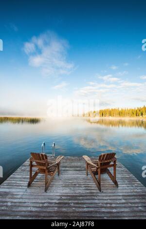 Sedie in legno sul molo che si affacciano sul lago all'alba Foto Stock