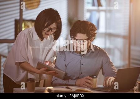 Asian giovane imprenditrice puntando alla tavoletta digitale e mostrando qualcosa al suo collega mentre si lavora sul computer portatile in ufficio Foto Stock