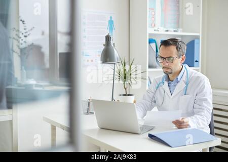 Grave medico maturo in occhiali e in camice bianco a lavorare con documenti e guardando il monitor del portatile mentre è seduto al suo posto di lavoro in ufficio Foto Stock
