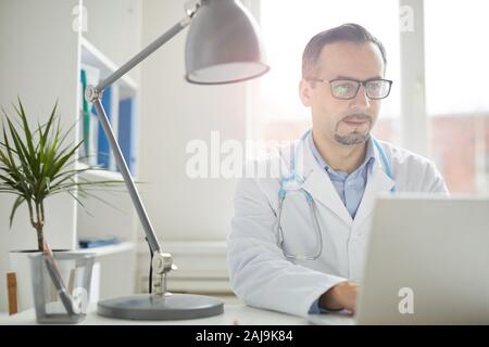 Maschio maturo medico in occhiali concentrando sulla sua opera egli lavora su computer portatile al tavolo in ufficio Foto Stock