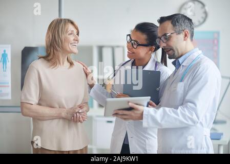 Maschio di medico e infermiere con tavoletta digitale e discutendo con sorridente donna matura il suo trattamento mentre si sta in piedi in ufficio Foto Stock