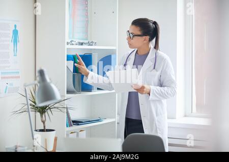 Medico donna in camice bianco in piedi vicino alla libreria e cercando le schede mediche dei suoi pazienti alla clinica medica Foto Stock