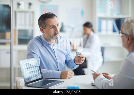 Uomo maturo tenendo una bottiglia di pillole e discutere il trattamento insieme con il suo medico al tavolo in ufficio Foto Stock