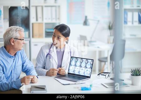 Giovani donne medico puntando al portatile con immagini a raggi x e discuterle con il senior paziente in ospedale Foto Stock
