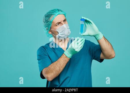 Medico chirurgo in frega e in abbigliamento protettivo tenuta tubo di prova con il liquido blu e esaminando contro lo sfondo blu Foto Stock