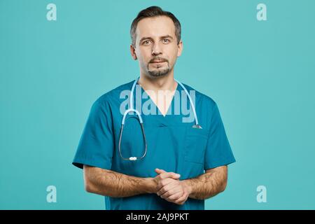 Ritratto di professional chirurgo maturo in scrubs guardando la fotocamera mentre in piedi contro lo sfondo blu Foto Stock