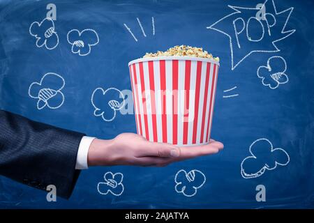 Maschio di mano che tiene il popcorn benna su sfondo blu. Industria cinematografica. La realizzazione del filmato. Arti cinematografiche Foto Stock