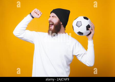 Barbuto uomo gioioso è urlando e celebrando, tenendo un pallone da calcio sulla parete gialla. Foto Stock