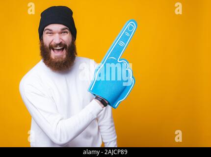 Allegro giovane uomo barbuto è ridere e puntando su una copia spazio con un grande ventilatore guanto sulla parete gialla. Foto Stock