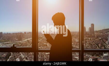 Silhouette di un giovane imprenditore di successo in tuta di office è la navigazione mobile phone, alla luce del sole. Egli è in piedi vicino alla finestra con vista panoramica della cittã nel suo ufficio presso il giorno lavorativo. Foto Stock