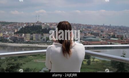 Brunette donna in maglia bianco guarda attraverso il binocolo esplorare cityscape stretching lungo la baia vista posteriore Foto Stock