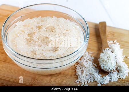 Riso ammollato, grano, acqua liquida torbida in ciotola di vetro su fondo di legno Foto Stock