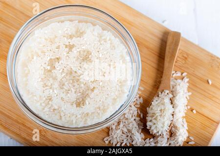 Riso ammollato, grano, acqua liquida torbida in ciotola di vetro su fondo di legno Foto Stock