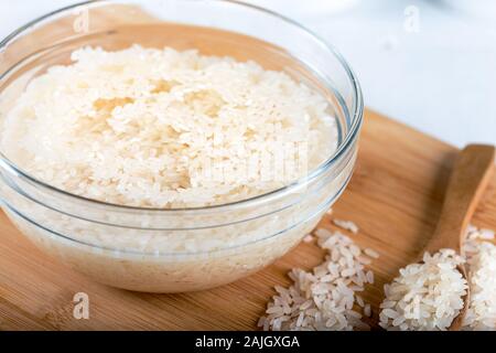 Riso ammollato, grano, acqua liquida torbida in ciotola di vetro su fondo di legno Foto Stock