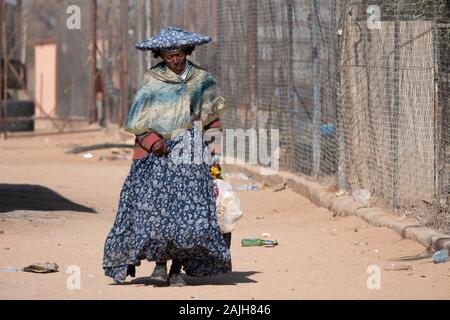 Omatjette, Regione di Erongo, Namibia - 24 Luglio 2019: Povero vecchio Herero donna in abito tradizionale con Horn-Shaped Hat a piedi attraverso una sporca Neighborhoo Foto Stock