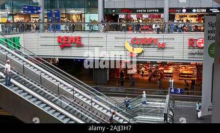 Colorati negozi su due livelli con scale mobili all'interno della moderna stazione ferroviaria principale di Berlino, Germania. Negozio di alimentari Rewe, e Curry 36 fast food. Foto Stock