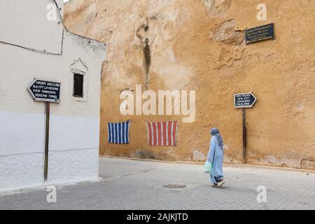 Donna in abito blu e con velo sulla testa, segnaletica che mostra il percorso per le attrazioni turistiche e tessuti appesi al muro, Fes (Fez), Marocco Foto Stock