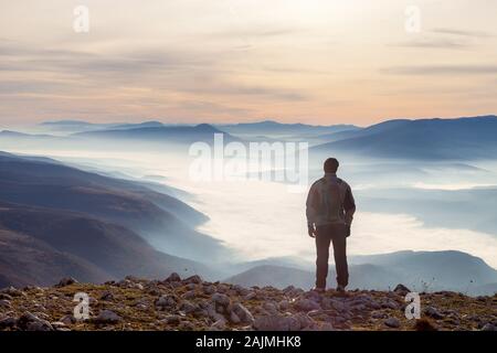 Escursionista di montagna che si erge ai margini della cima della montagna e guarda una valle piena di nebbia fitta durante l'alba colorata Foto Stock