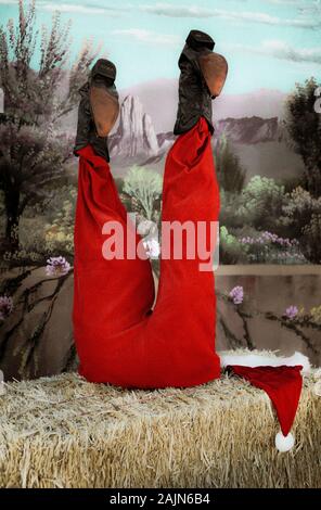 Una buffa set fotografico per ritratti con un capovolto nella metà inferiore di Santa Claus in rosso vestito con stivali da cowboy contro un deserto dipinto sullo sfondo Foto Stock