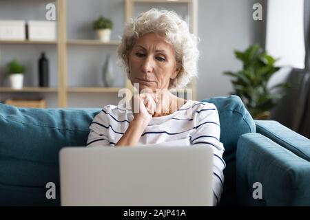 Vecchia donna accomodarsi all'interno guarda lo schermo del notebook si sente confusa Foto Stock