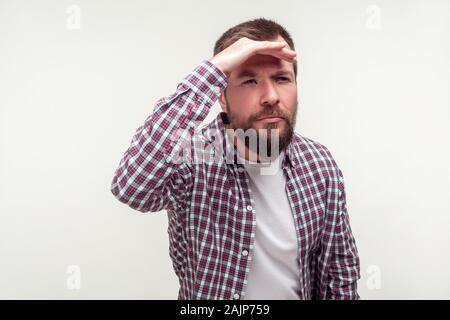 Ritratto di calma attento uomo barbuto in casuale plaid shirt in piedi tenendo la mano sopra gli occhi e guardare lontano, guardando un orizzonte a lunga distanza. Foto Stock