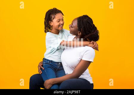 Felice famiglia amorevole. Overjoyed figlia nera seduto sulla madre di giro guardando ogni altro su sfondo giallo Foto Stock