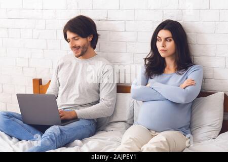 Donna incinta offeso da suo marito che giocare con i videogame sul computer portatile e non dando attenzione mentre sono seduti sul letto insieme, spazio di copia Foto Stock