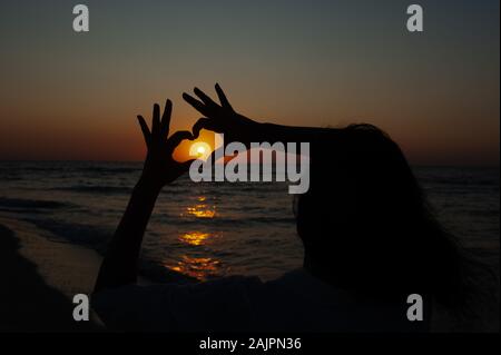 Segno di cuore con le mani contro il tramonto e l'alba sul mare Foto Stock