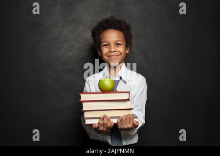 Felice bambino nero studente contenere libri e apple e sorridente su sfondo lavagna Foto Stock