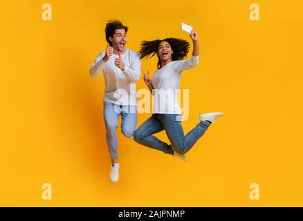 Gioiosa interracial giovane tenendo selfie durante il salto, mostrando pollice in alto e i gesti di pace, per divertirsi insieme su sfondo giallo in studio Foto Stock