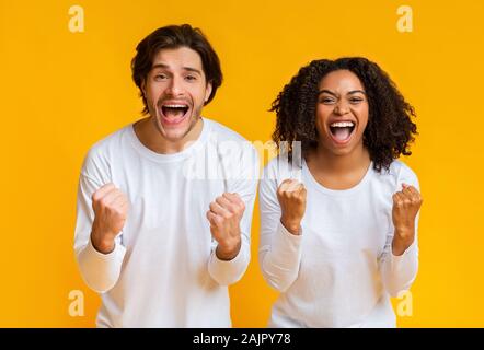 Sì. Felice interracial giovane urlando e stringendo i pugni con entusiasmo, tifo e posa insieme su giallo di sfondo per studio con spazio libero Foto Stock