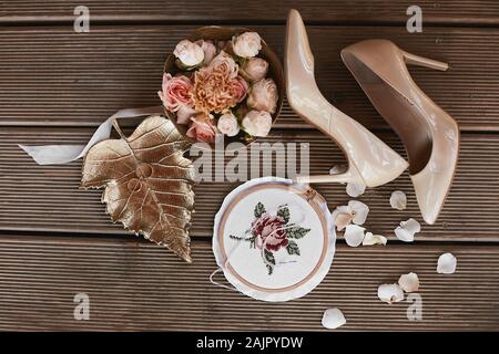 Rose e peonie fiori in una scatola rotonda, gli anelli di nozze sulla foglia oro, bridal scarpe sui tacchi alti su sfondo di legno, vista dall'alto. Dettagli Nozze Foto Stock