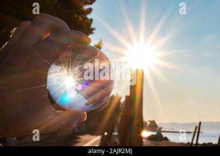 Mano che tiene un bicchiere di cristallo sfera, mattino dura sole nel cielo si riflette nella sfera. Contorni di spiaggia in background Foto Stock