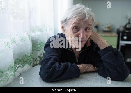 Lonely vecchia donna seduta in cucina dalla finestra della sua casa. Foto Stock