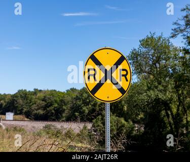 Giallo attraversamento ferroviario segno di avvertimento sulla strada rurale con alberi e binari del treno in background Foto Stock