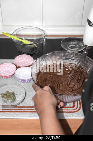Persona prepara deliziosi muffin al cioccolato con boccioli di marijuana a casa. Sbattere l'impasto di cioccolato con la miscela di ingredienti. Foto Stock
