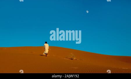 Un marocchino uomo che indossa a lunghezza piena tradizionale abbigliamento nomade, passeggiate a piedi nudi in salita verso il crinale di un rosso dune di sabbia nel deserto del Sahara. Foto Stock