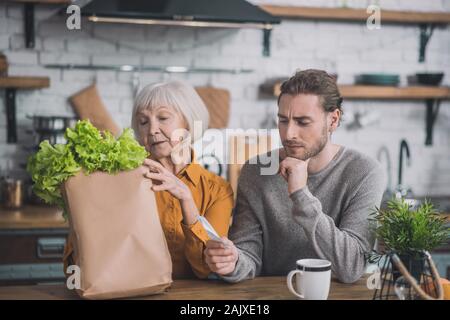 Grey-haired mom e suo figlio seduto in cucina dopo lo shopping Foto Stock