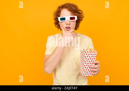 I capelli rossi bello teen boy in T-shirt con bicchieri di film con popcorn nelle sue mani su sfondo giallo con spazio di copia Foto Stock