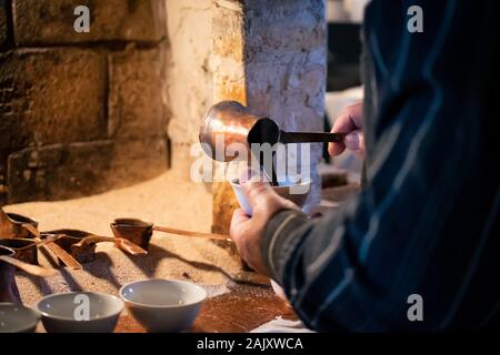 L'uomo versando il caffè turco a partire da un tradizionale pentola di ottone, preparato sulla sabbia a caldo in un forno Foto Stock