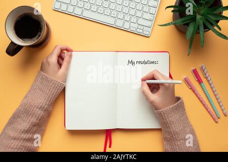 Stock Foto di una giovane donna le mani la scrittura in un 2020 anno nuovo notebook con elenco di risoluzioni e oggetti su sfondo giallo Foto Stock