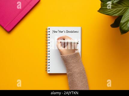 Stock Foto di una giovane donna la scrittura a mano in un 2020 anno nuovo notebook con elenco di risoluzioni e oggetti su sfondo giallo Foto Stock